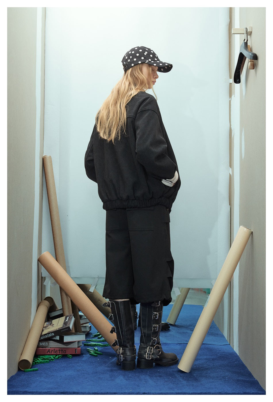 Model shown from behind in an oversized black jacket, loose black pants, buckle boots, and a black cap with white dots.