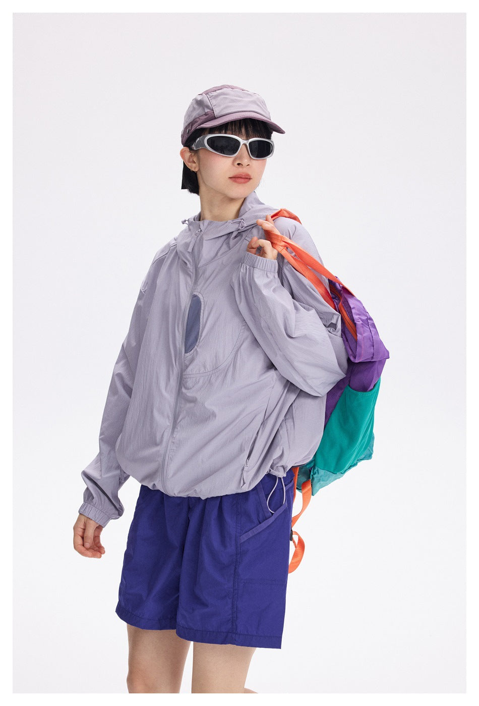 Closer front view of a model wearing a light lavender zip-up hooded jacket, royal blue shorts, a cap and sunglasses.