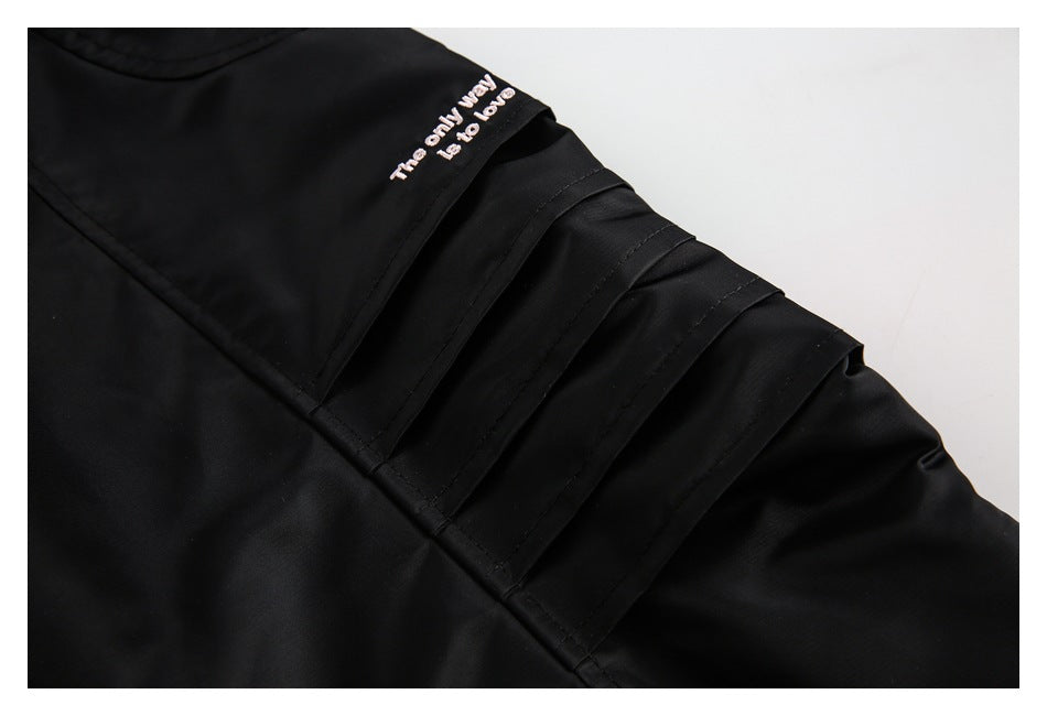 Close-up of black jacket sleeve with layered stitched panels and a small pale pink embroidered slogan.