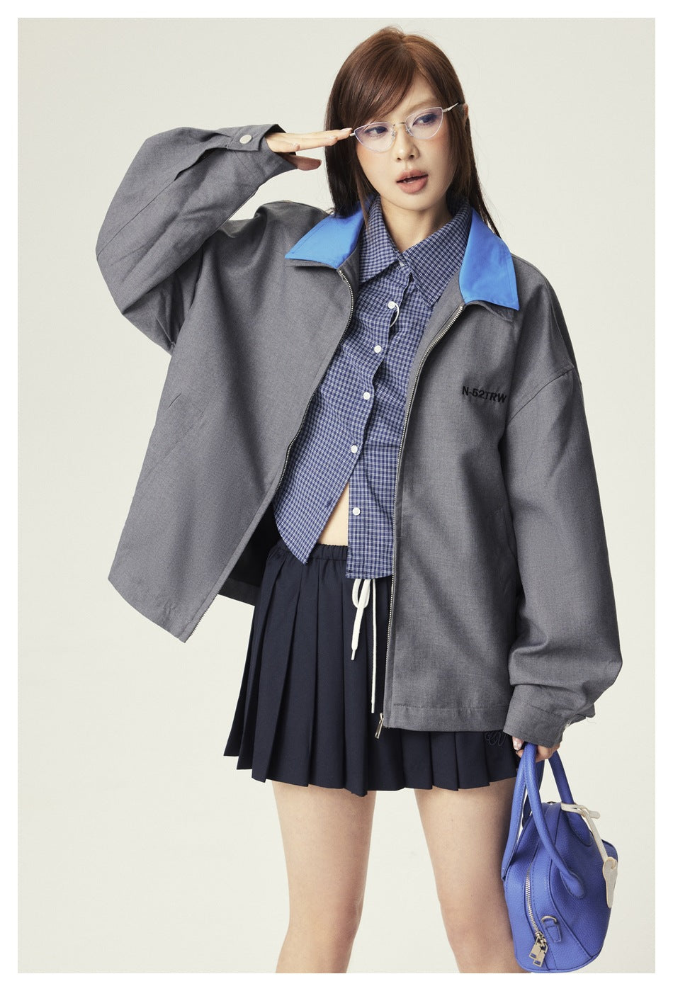Model in a gray oversized jacket with a blue collar, shown with the front open over a checked shirt and pleated skirt.