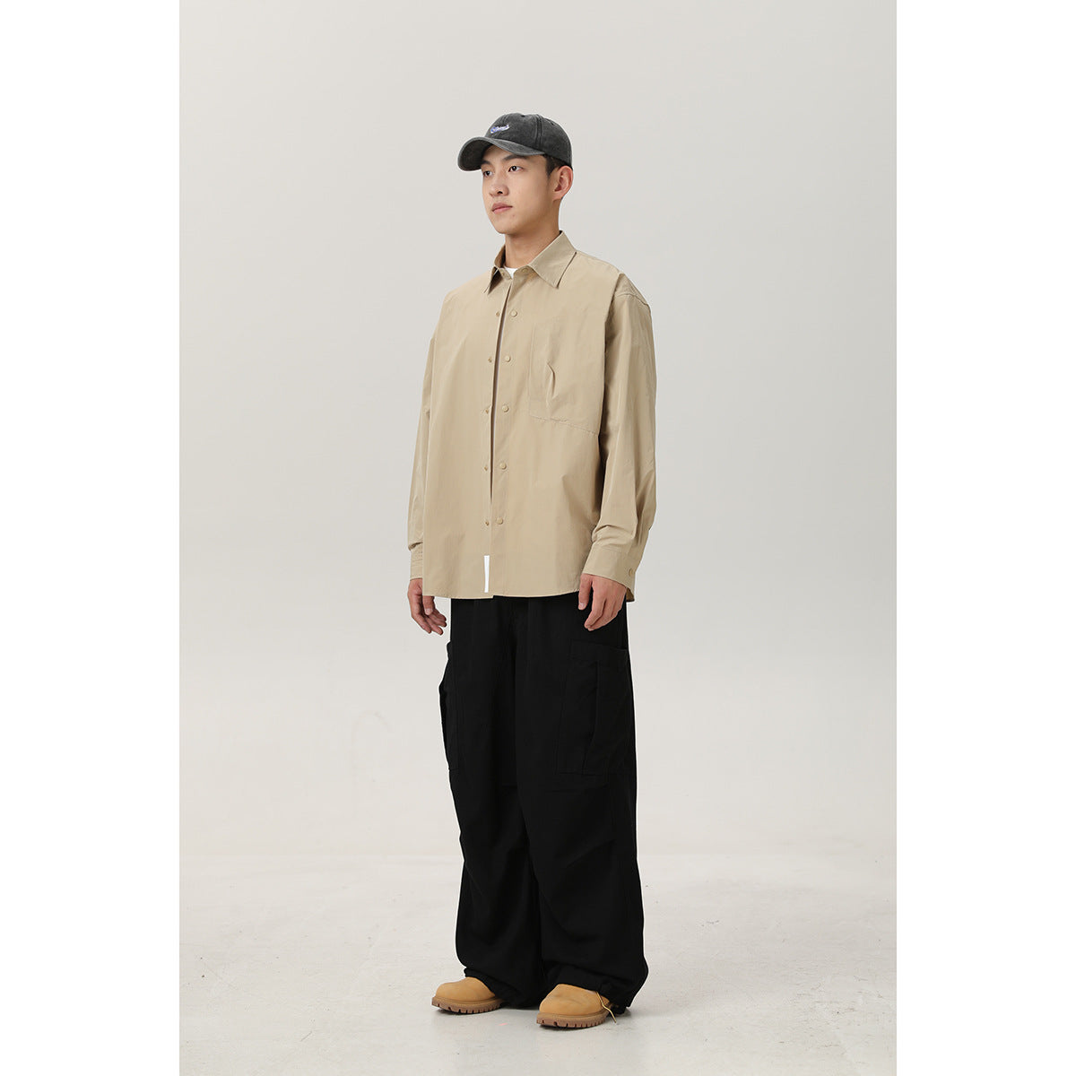Model wearing a beige snap-front shirt with chest pocket, loose black cargo pants, tan shoes, and a dark cap.