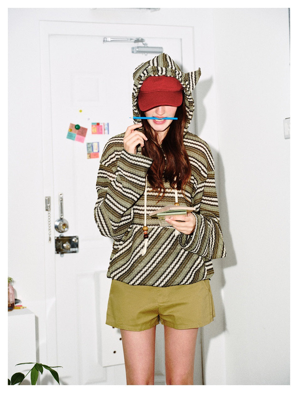 Model wearing the green striped cat-ear hoodie with a red cap and olive shorts, holding a pencil and notepad