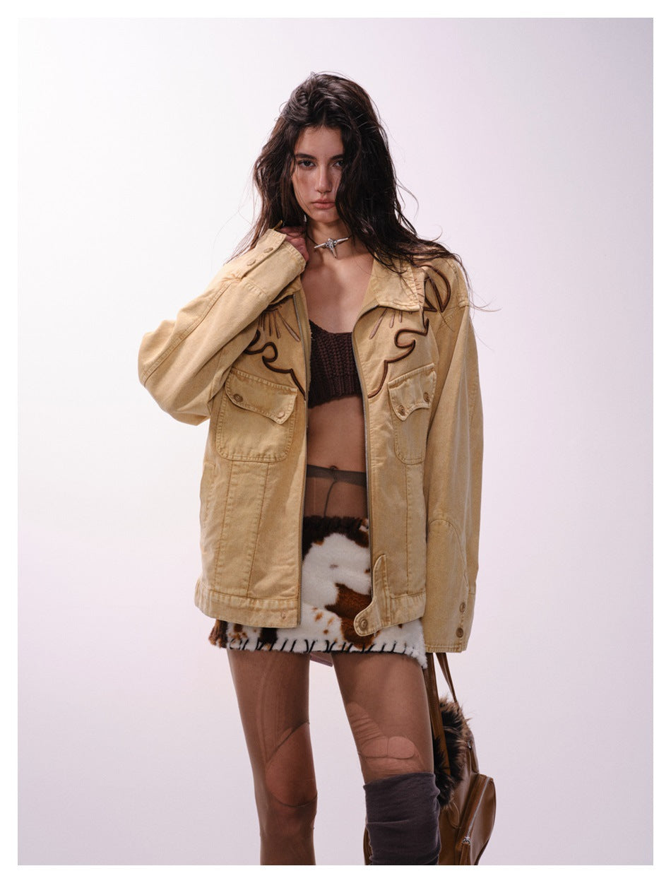 Model wearing an open tan jacket with dark embroidered motifs and flap pockets, styled with a black crop top and patterned
