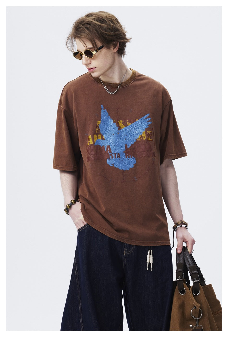 Close-up of model wearing brown T-shirt with textured blue bird graphic, sunglasses, chain necklace and dark jeans