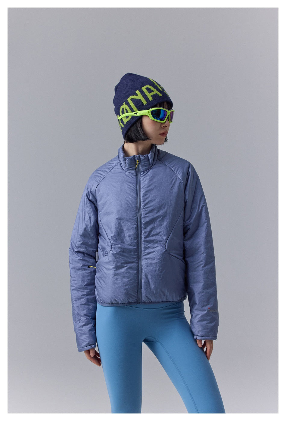 Front view of a model in a periwinkle-blue padded zip jacket and blue leggings, wearing a navy beanie and neon sunglasses.