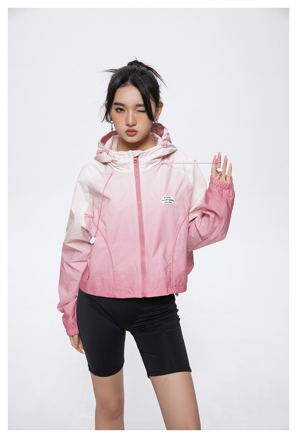 Model wearing a pale-to-rose pink hooded zip jacket with drawcords and elastic cuffs, paired with black shorts.