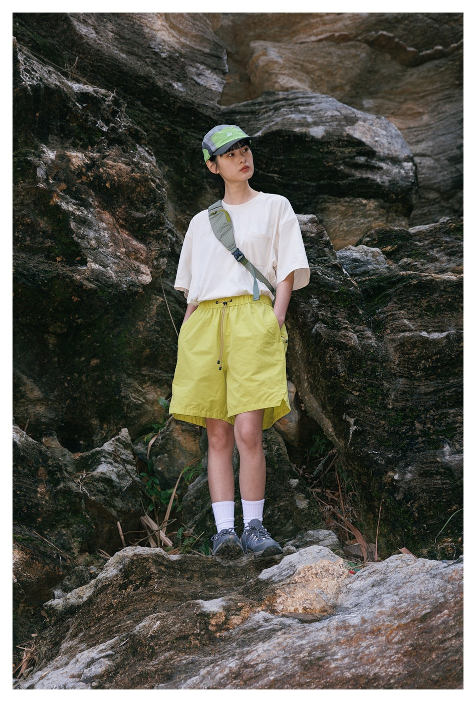 Model in a cream pocket T-shirt with yellow shorts, cap and crossbody bag, hands in pockets, on rocks