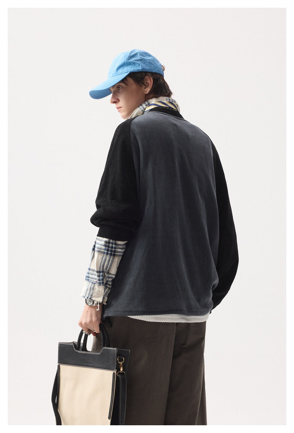 Rear view of a model wearing a charcoal raglan sweater over a plaid shirt, with a blue cap and carrying a tote.