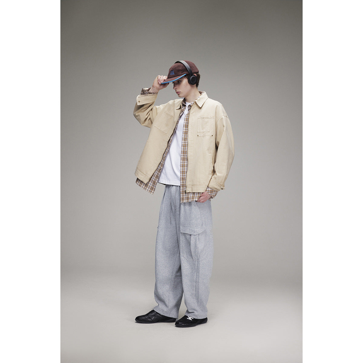 Model wearing a beige utility jacket over a plaid shirt and white tee, with loose gray pants and a brown cap.