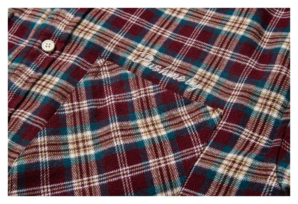 Close-up of burgundy, teal, cream, and brown plaid shirt fabric with a white button, chest pocket, and cursive embroidery.