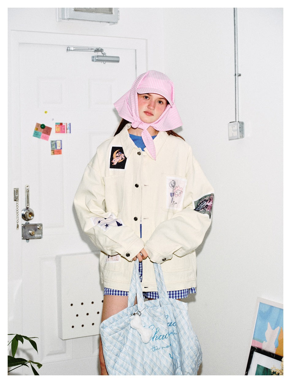 Model wearing an ivory illustrated-patch jacket with blue gingham shorts and a pink striped bonnet, holding a quilted blue