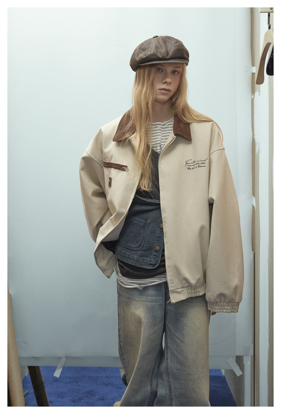 Model wearing a cream zip jacket with a brown collar and trim, styled with jeans and a brown cap.