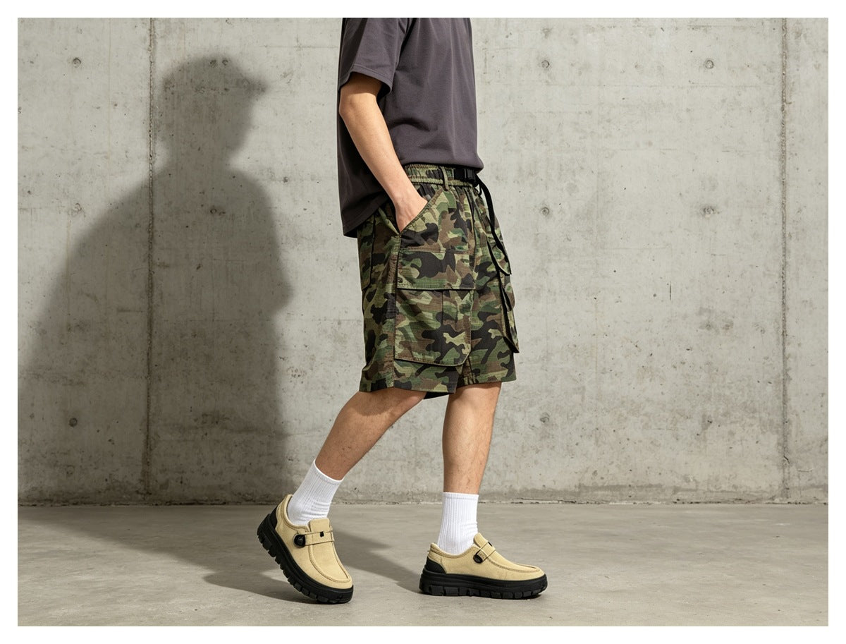 Person wearing camouflage cargo shorts with a gray tee, white socks and tan shoes, side view with hand in pocket