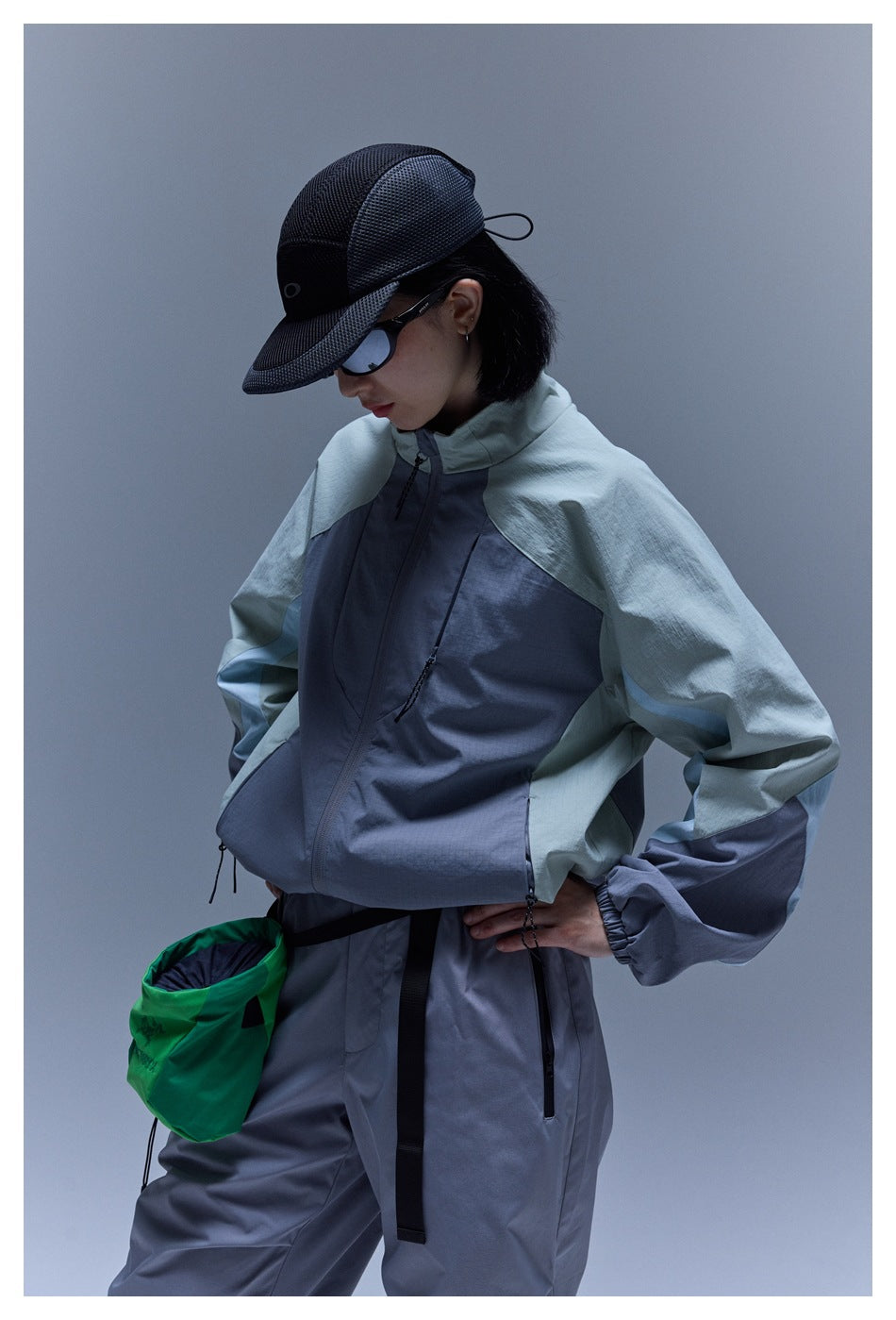 Model wearing a slate-gray and pale sage color-block jacket with matching pants, a cap, sunglasses, and a green pouch.