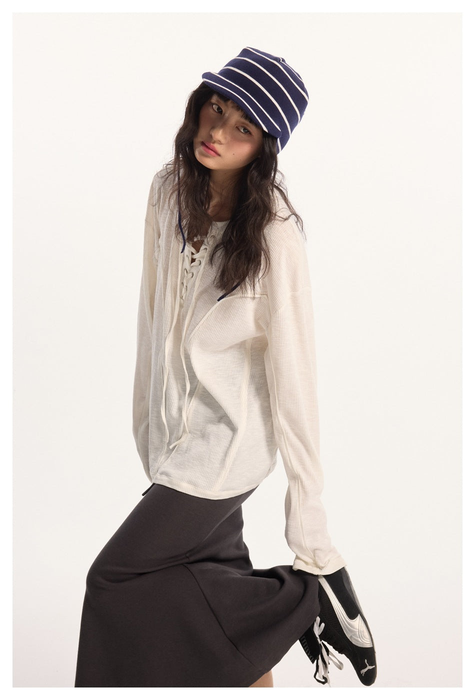 Model wearing an ivory oversized lace-up top, dark skirt, and navy striped cap in a seated pose.