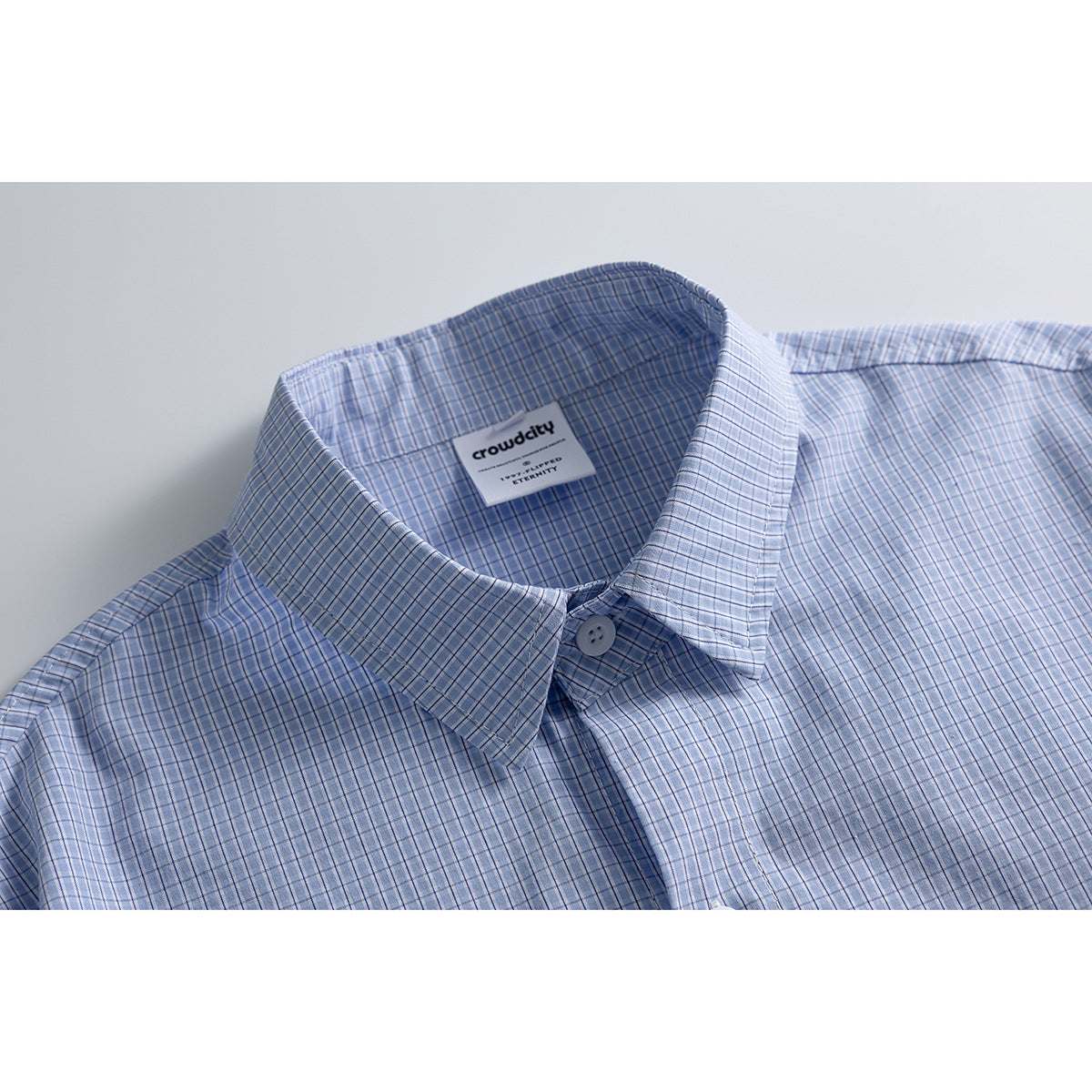 Close-up of a light blue checked shirt collar and button, with a woven brand label inside the neck.