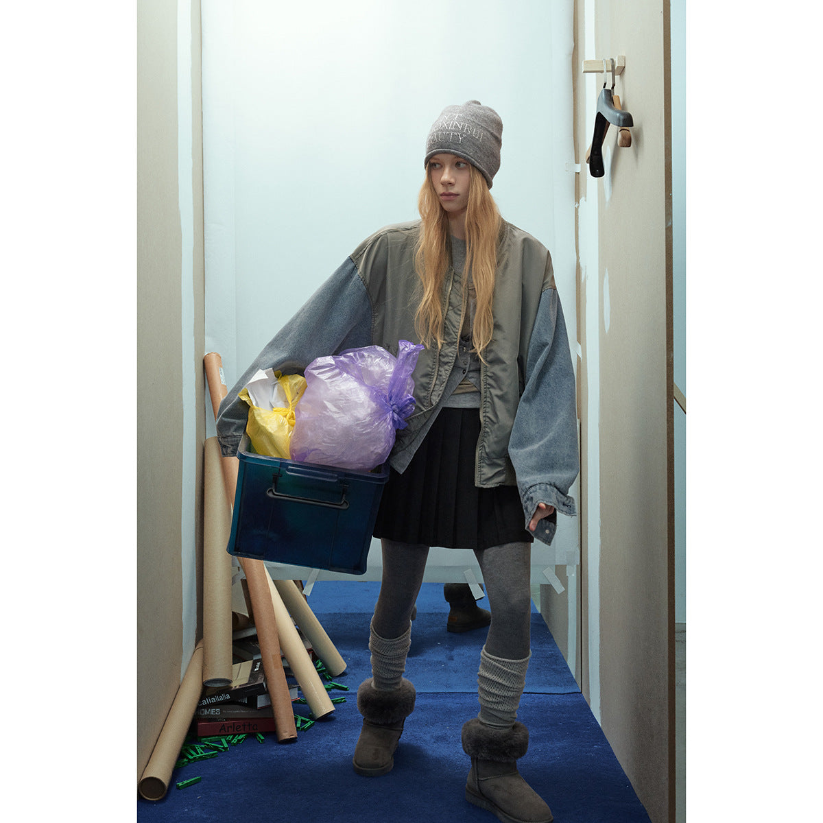 Model wearing an oversized gray-green bomber jacket with blue denim sleeves, a gray beanie, black skirt and gray leggings.