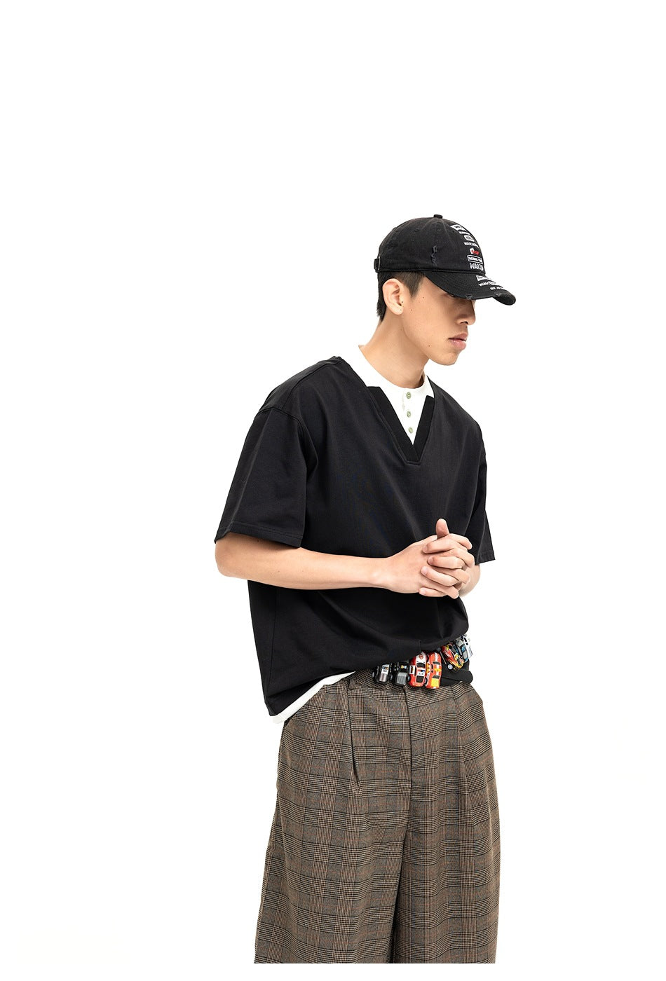 Model wearing black V-neck tee with white collar and button placket, plaid pants and graphic cap, three-quarter view