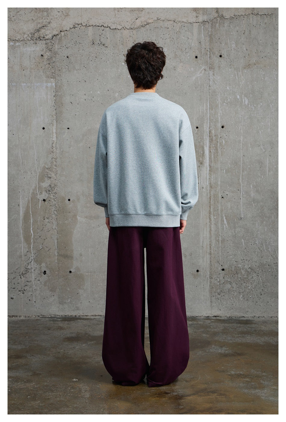 Rear view of a model wearing a light gray crewneck sweatshirt and loose burgundy trousers.