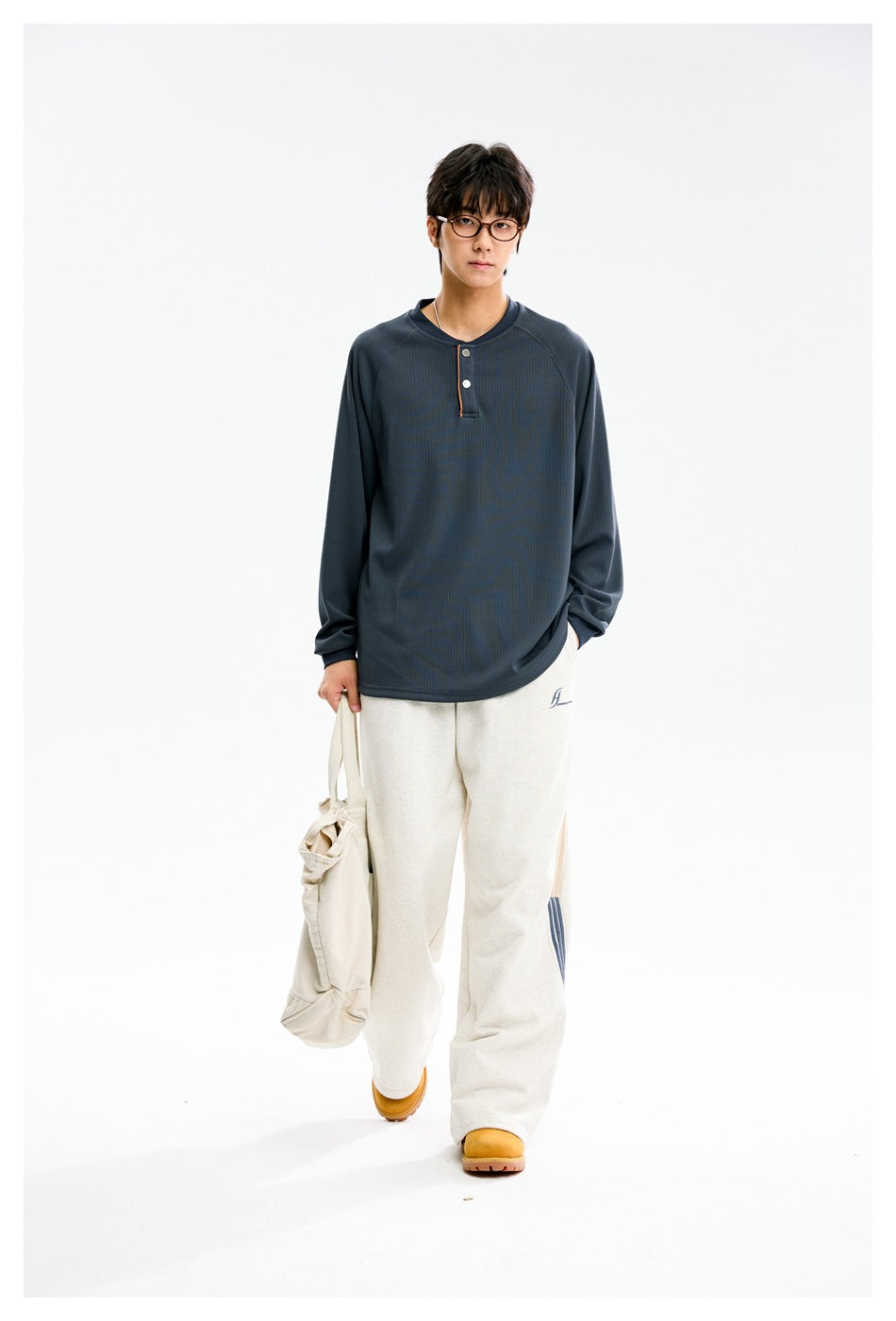 Model wearing a slate blue long-sleeve henley with white buttons and loose cream pants, carrying a canvas tote.