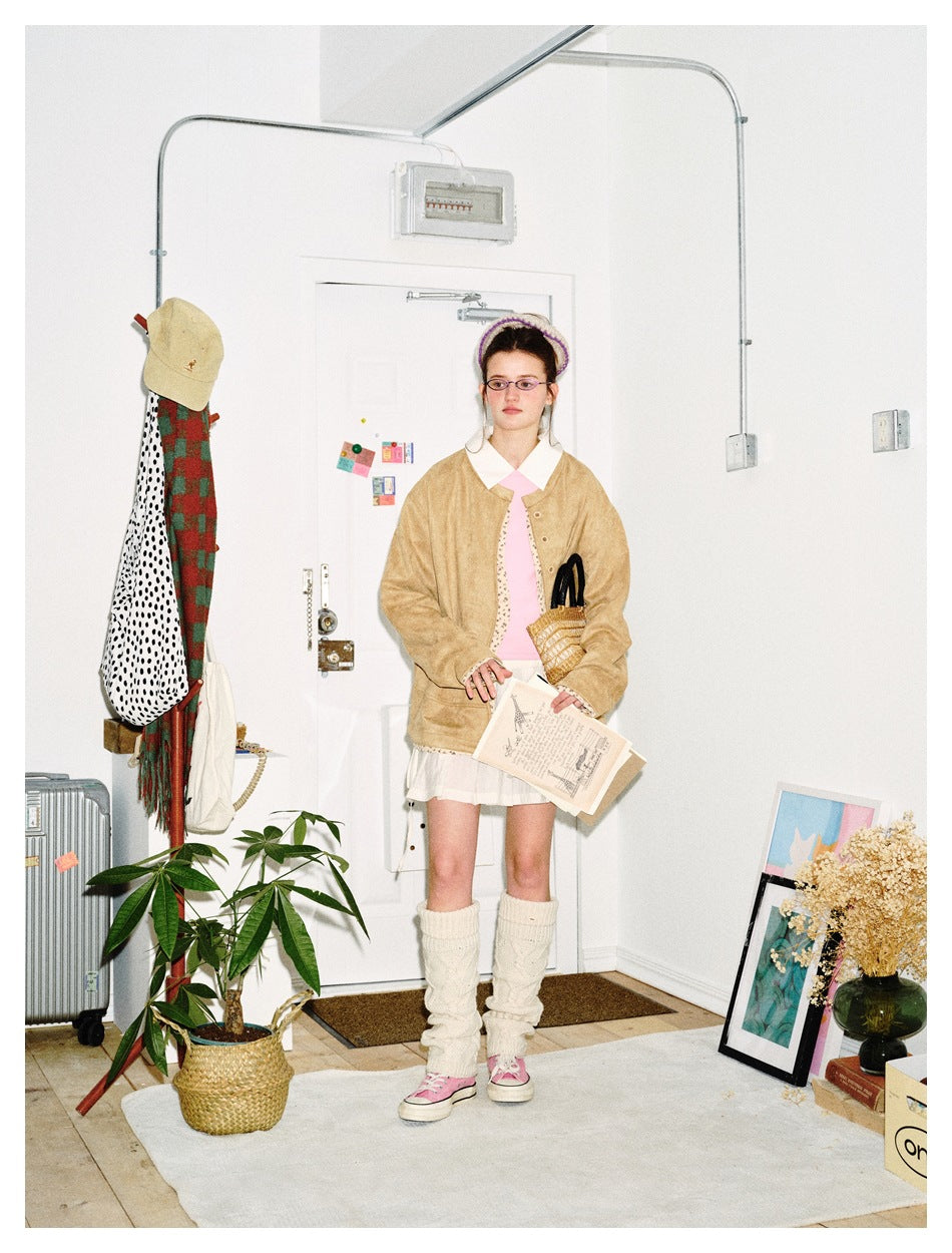 Model wearing camel suede-look jacket open over a pink top and white pleated skirt in a bright room