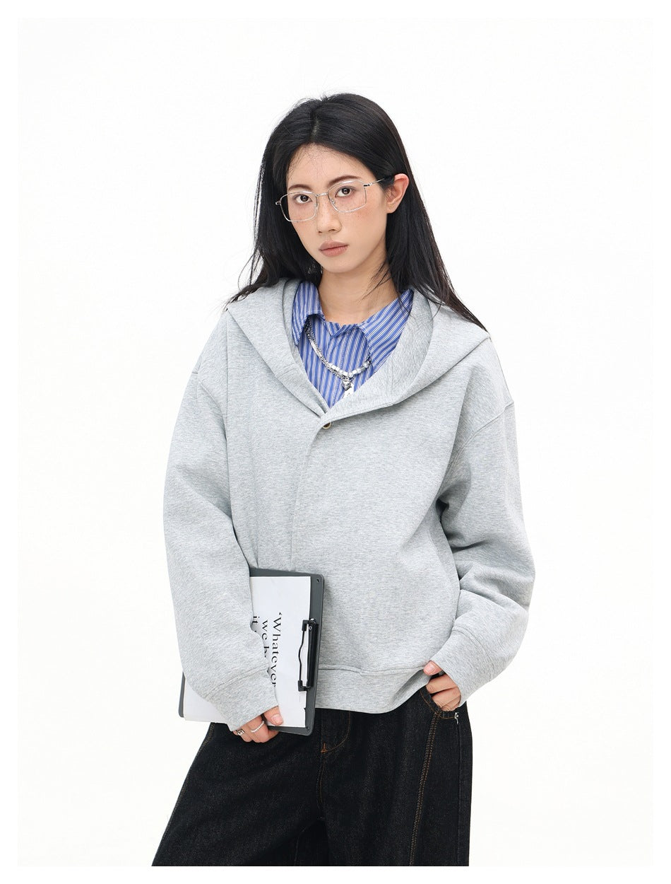 Model wearing a light gray oversized hooded sweatshirt with a wide folded collar and dark jeans, holding a clipboard.
