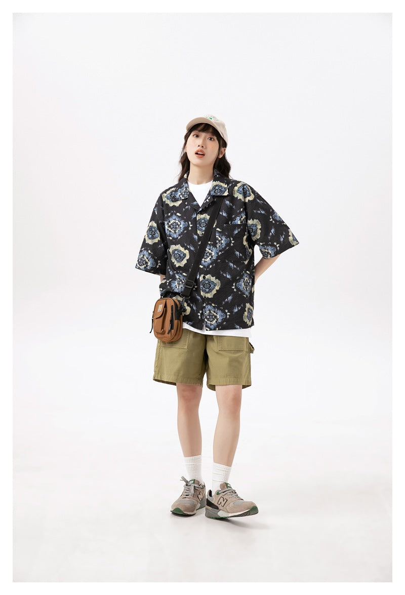 Model wearing a black short-sleeve shirt with blue and beige motifs, olive shorts, a cap, and a crossbody bag.