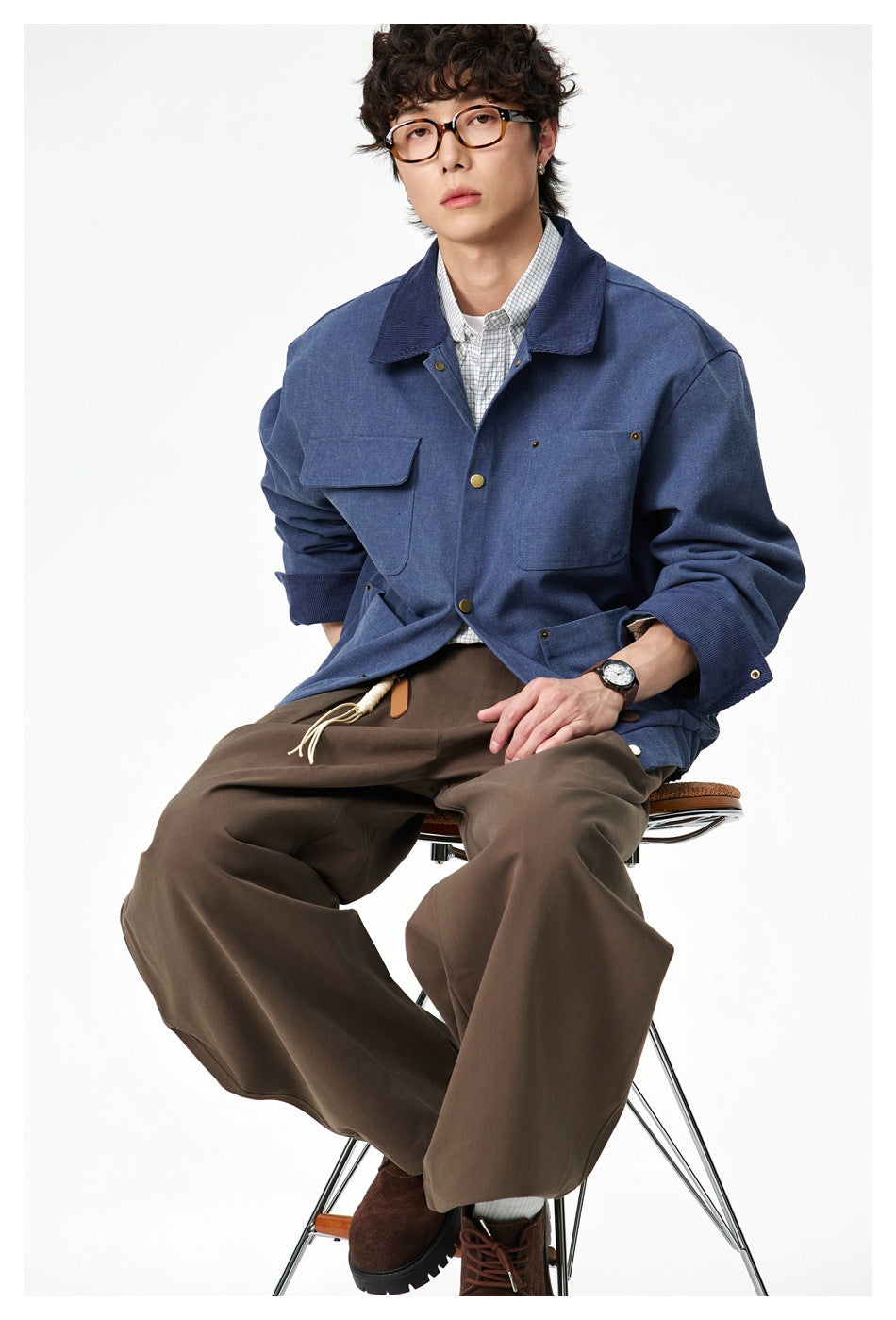 Man seated on a stool wearing the blue snap-front jacket with a plaid shirt and wide brown pants