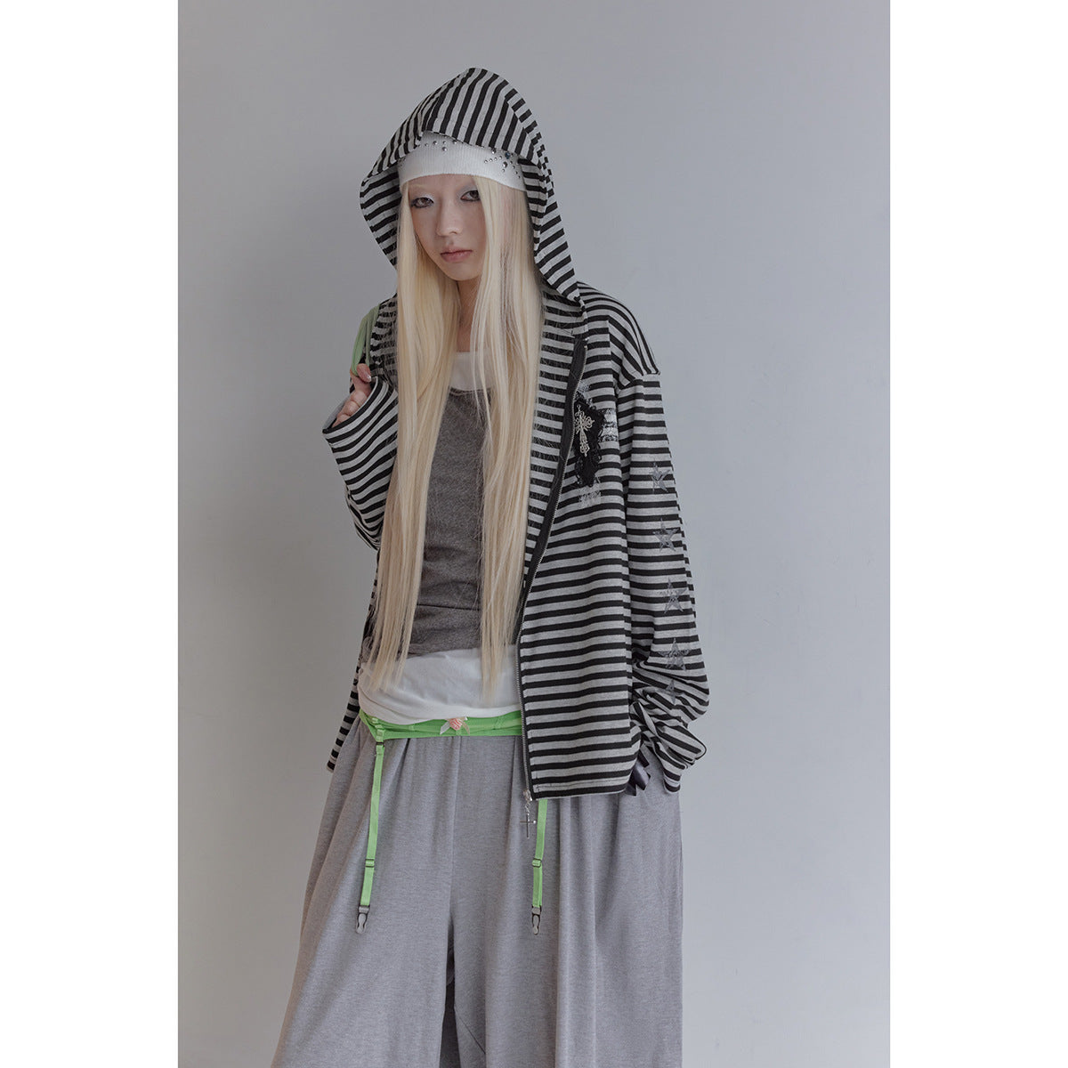 Model wearing an open black-and-gray striped hooded zip jacket with a cross patch and loose gray pants.