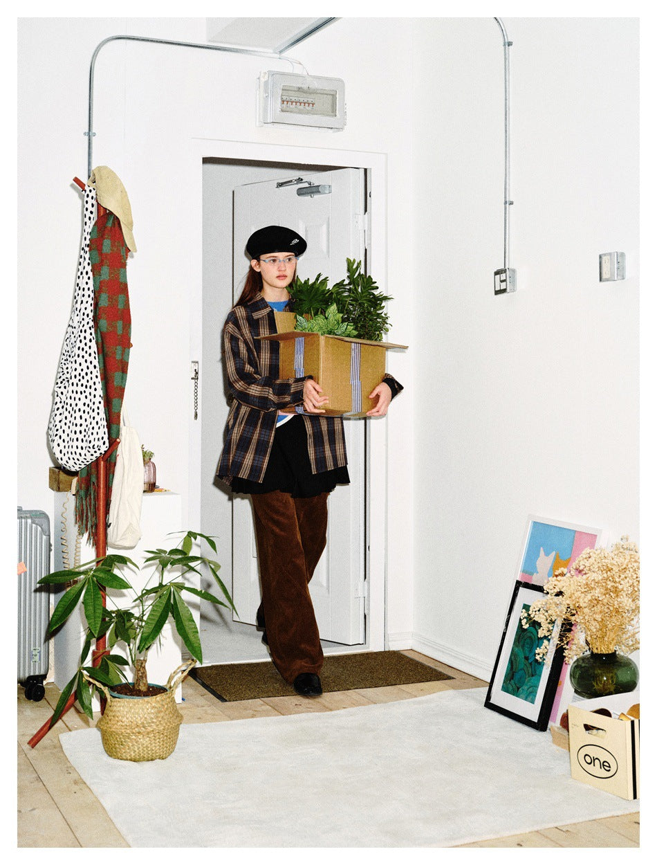 Model wearing a blue, brown and beige plaid shirt and brown pants while carrying a box of plants.