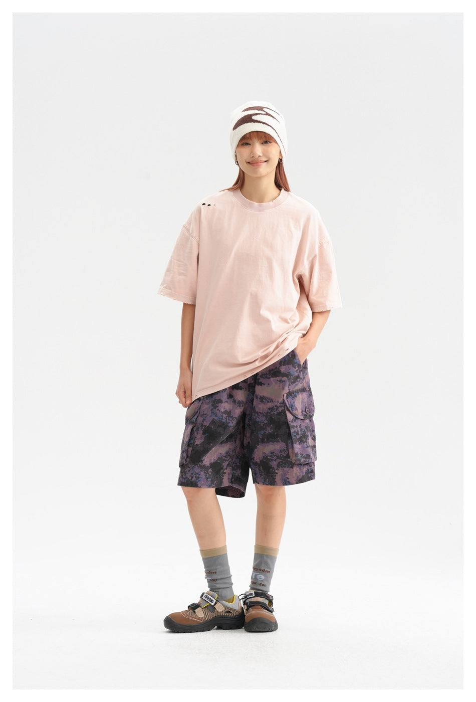 Full-length model wearing an oversized pale pink T-shirt, purple camouflage cargo shorts, and a white patterned beanie.