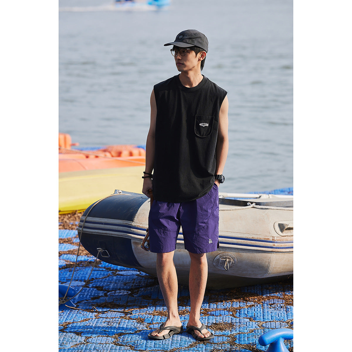 Full-length view of a model wearing a black sleeveless pocket shirt and purple shorts beside a boat.