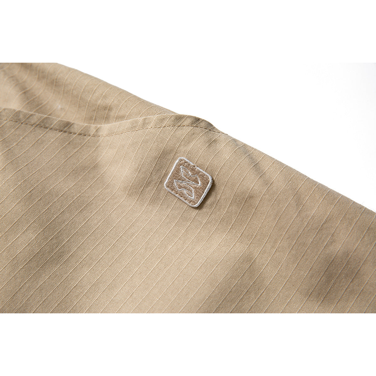 Close-up of tan shorts fabric with a small stitched emblem near a curved seam.