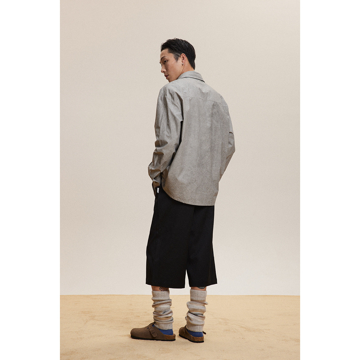 Model viewed from behind wearing a loose gray button-front shirt with black shorts and slouchy socks.