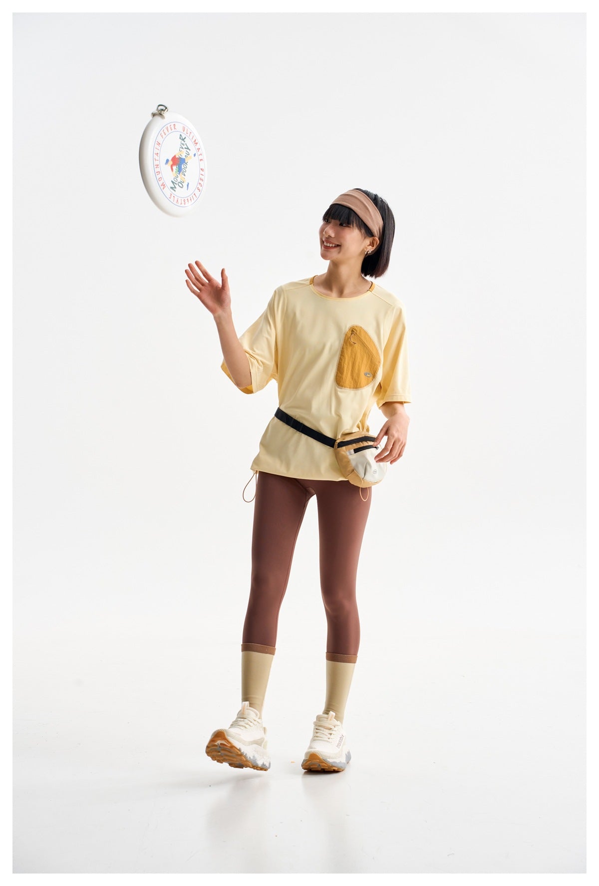 Model in a pale yellow oversized tee with a tan chest pocket, brown leggings, cream gaiters and a waist pouch.