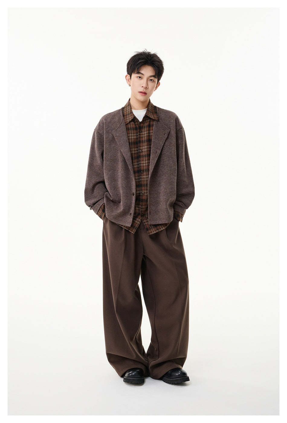 Model wears a loose brown button-front jacket with wide lapels over a brown plaid shirt and wide-leg trousers.