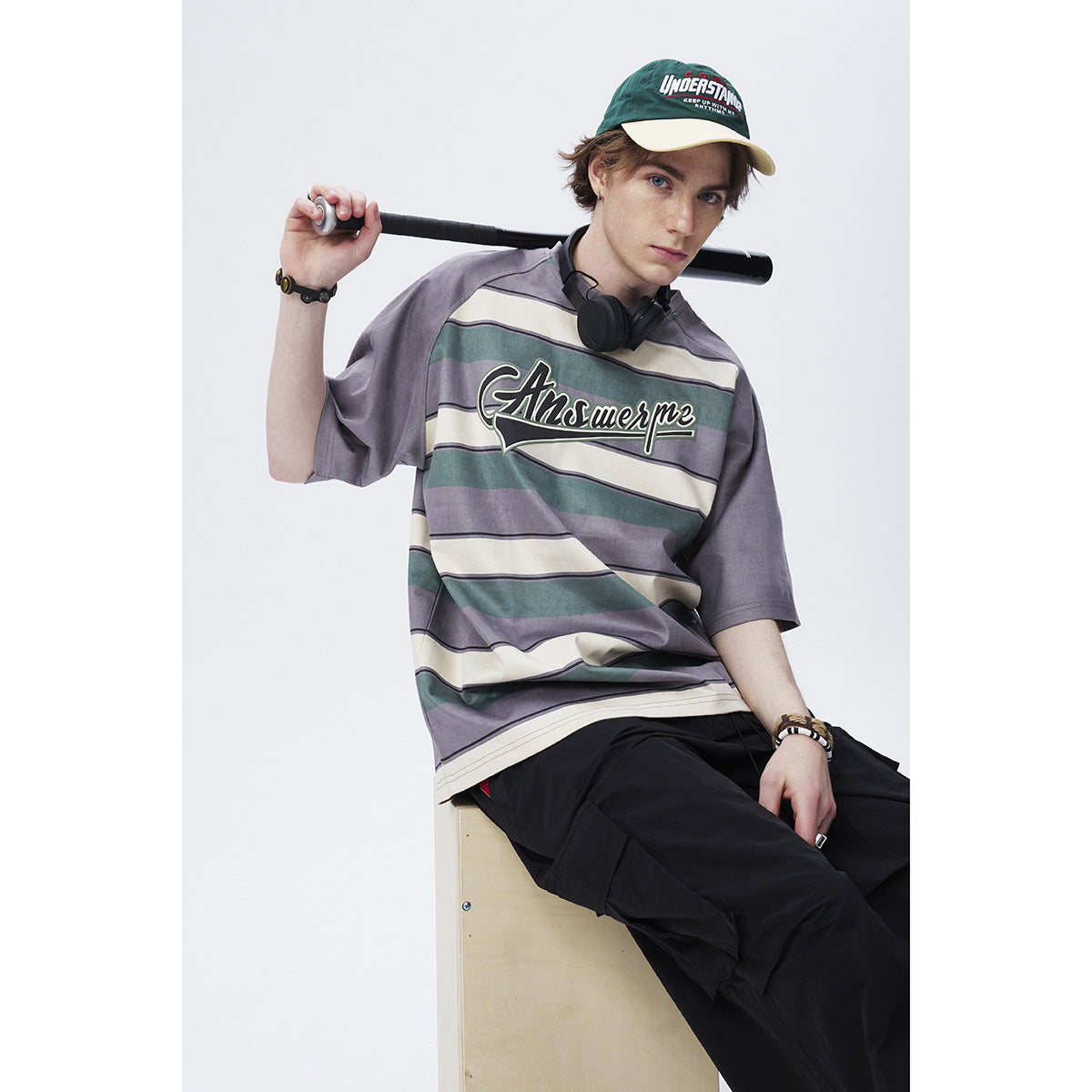 Seated model wearing an oversized gray, teal, and cream striped T-shirt with chest lettering and black trousers.