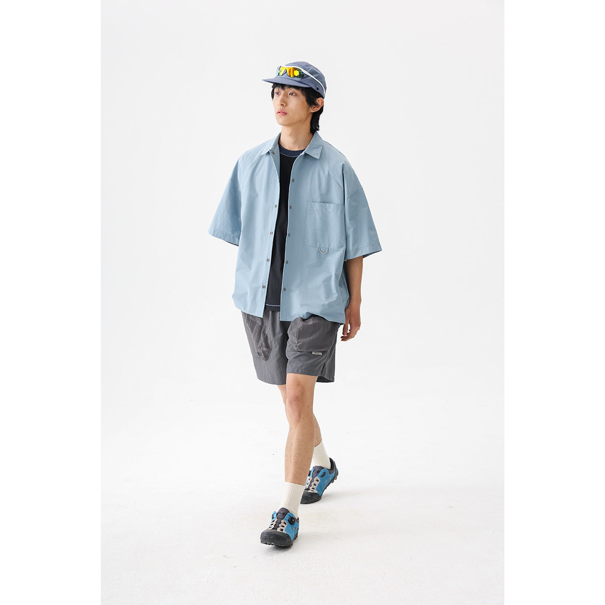 Model wearing a pale blue short-sleeve button-up shirt, dark gray cargo shorts, and a cap against a white studio background.