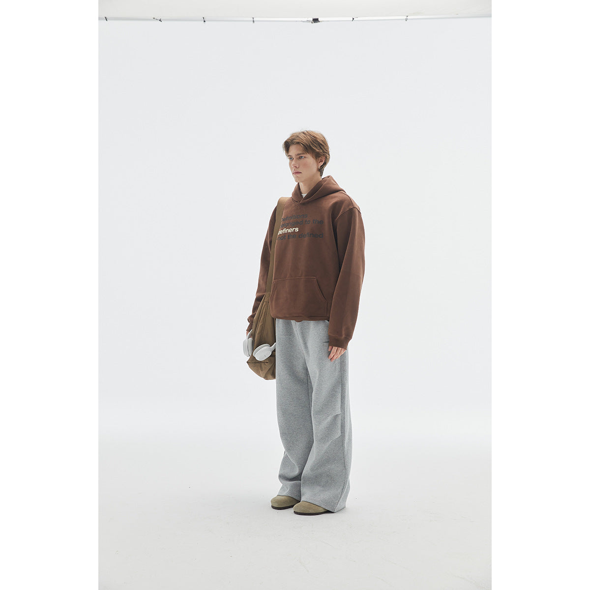 Model in a brown text-print hoodie and wide gray pants, shown at an angle with a shoulder bag and headphones.