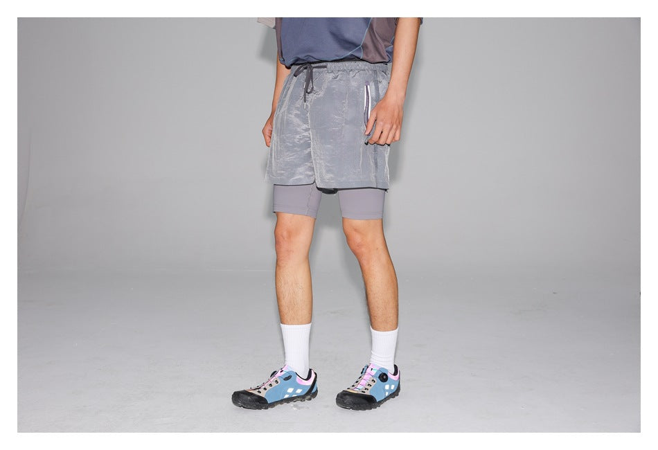 Cropped model wearing shiny silver-gray running shorts over fitted gray shorts, with white socks and blue trail shoes.