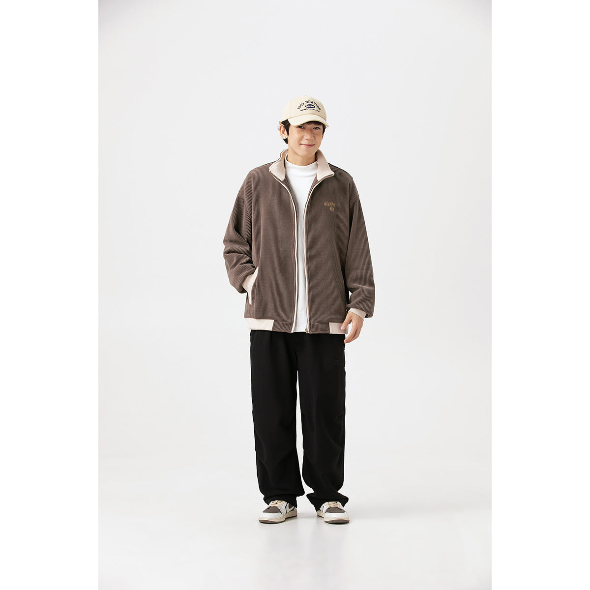 Full-length view of a model wearing a brown ribbed zip jacket with cream trim, black pants and sneakers.