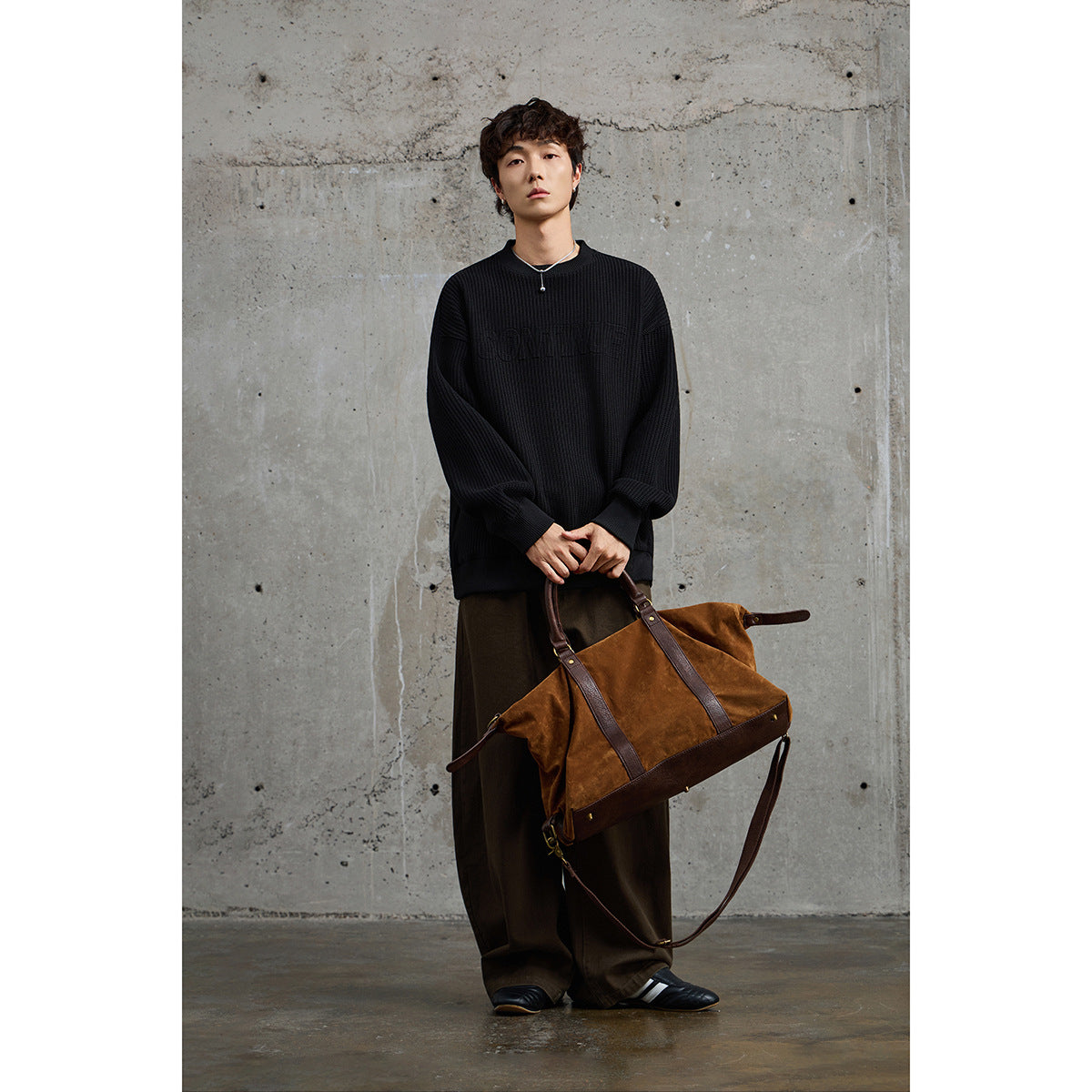 Model wearing a black textured crewneck sweater with dark trousers, holding a brown leather bag.