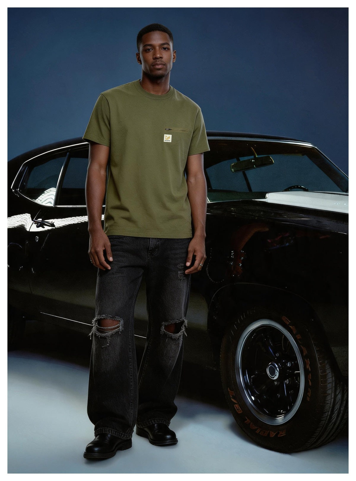 Man wearing an olive green short-sleeve T-shirt with zip chest pocket, black ripped jeans, standing by a black classic car