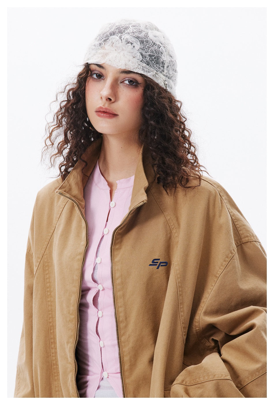 Close view of a model wearing a tan jacket, showing its zip front, panel seams and small embroidered chest emblem.