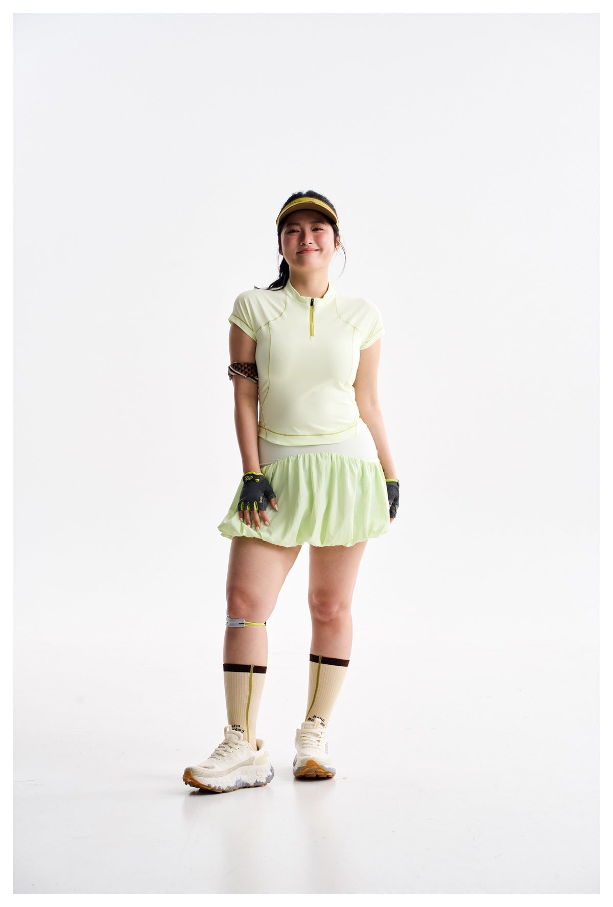 Model wearing a pale yellow-green zip-neck top and bubble skirt with a visor, gloves, cream socks, and sneakers.