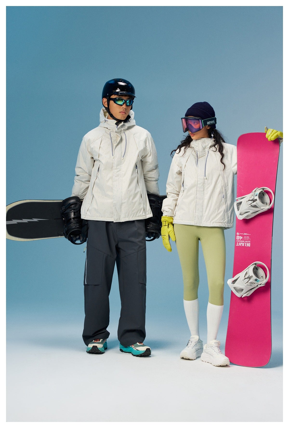 Man and woman wearing off-white hooded jackets with snowboards against a blue studio backdrop
