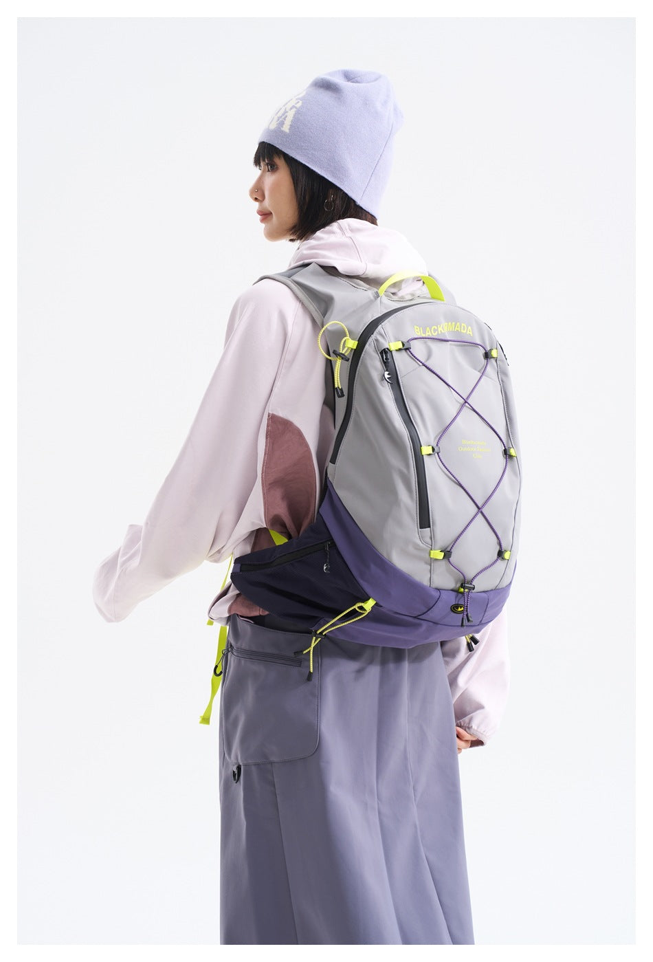 Rear view of a model wearing a gray and purple backpack with cord lacing, a pale jacket, and a gray skirt.
