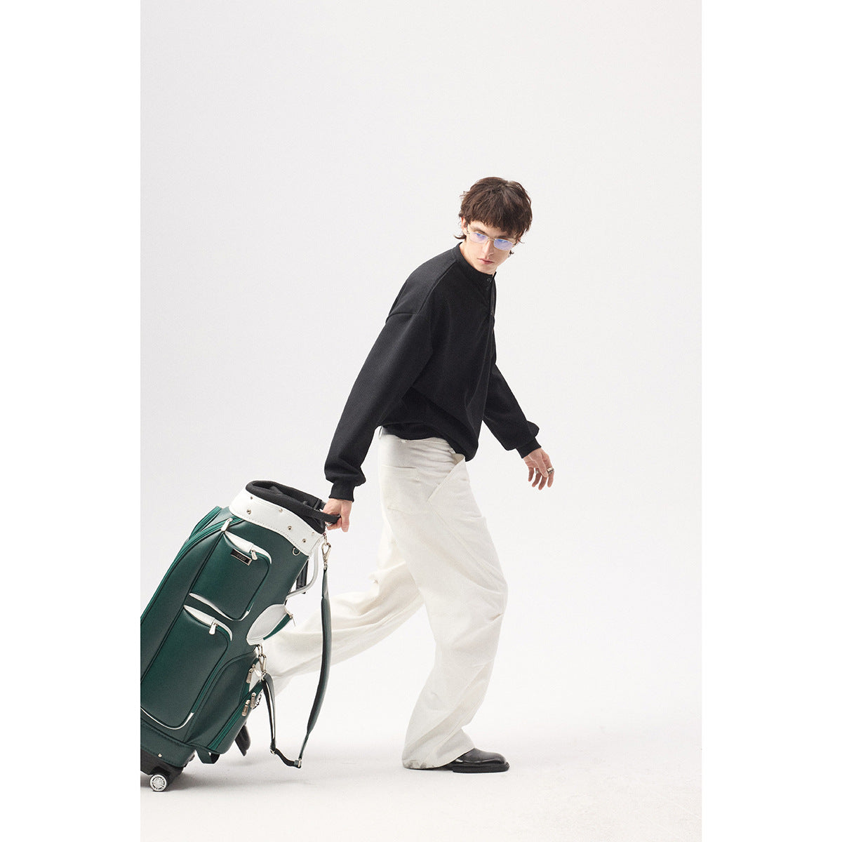 Model in profile wearing a black long-sleeve top and white trousers while pulling a golf bag.