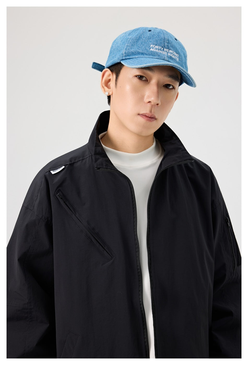 Close-up of a model in a black stand-collar jacket with diagonal chest zip pocket, white tee and blue denim cap