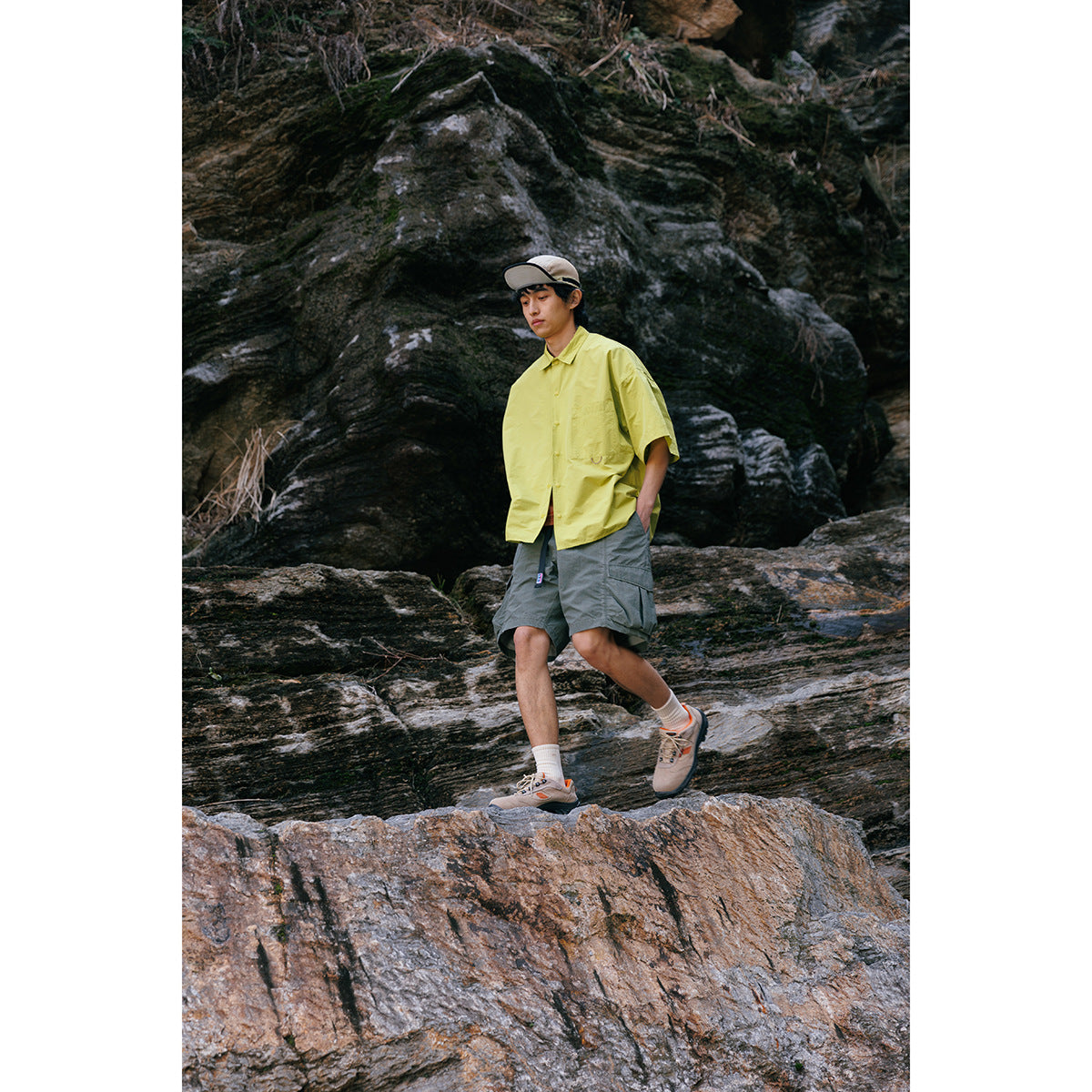 Model wearing a bright yellow-green short-sleeve shirt and gray cargo shorts while walking across rocky terrain.
